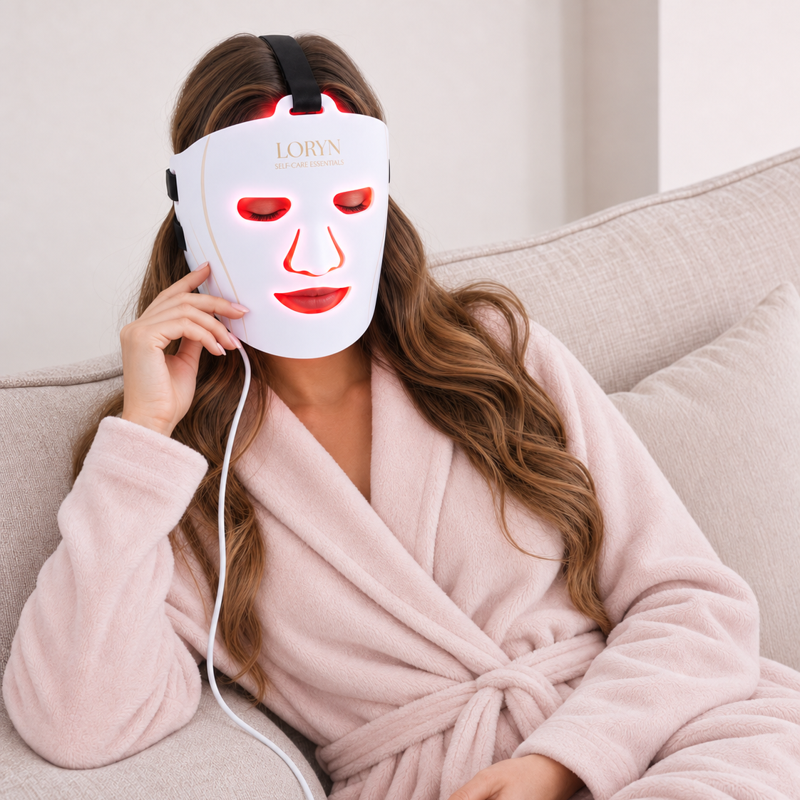 A woman in a pink robe sits on a beige sofa using a white LORYN LED light therapy face mask with glowing red lights and holding a control unit.
