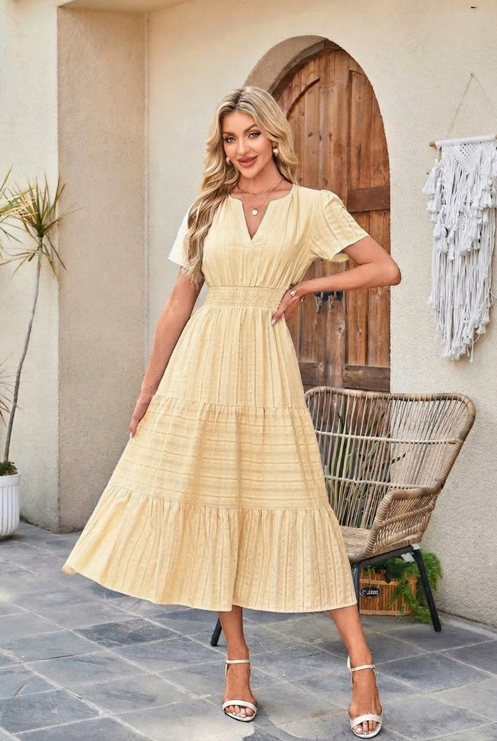 Woman in a beige dress standing in an outdoor setting with a wooden door and wicker chair.